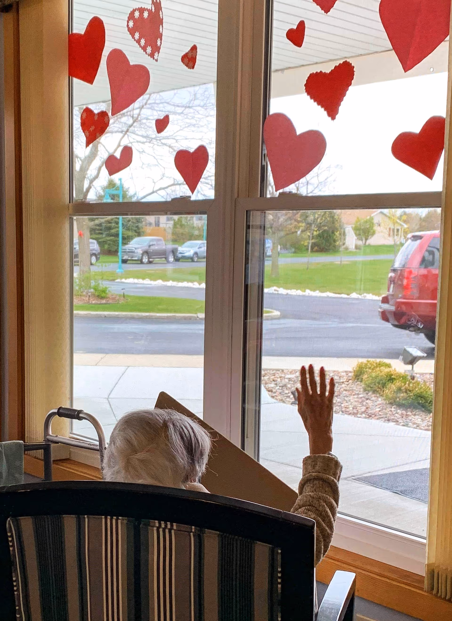 An elderly person with white hair sitting in a chair near a window decorated with red paper hearts. The person is raising their hand towards the window, and a walker is positioned nearby. Outside the window, a parking lot and some greenery are visible.