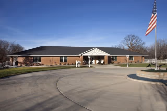 Single-story brick building with a black roof and white trim, featuring a covered entrance with white columns. There is a circular driveway in front and an American flag on a flagpole to the right. The sky is clear and blue, and there are leafless trees in the background.