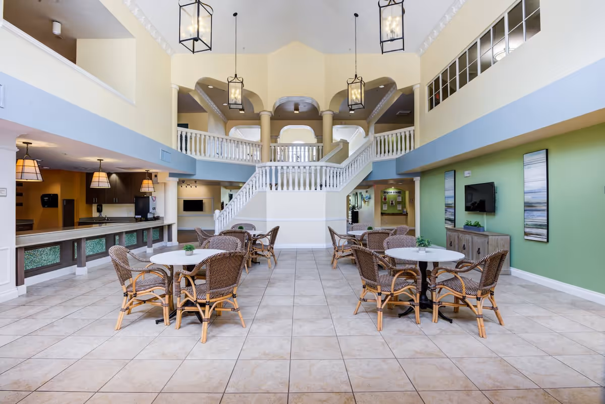Spacious interior common area with high ceilings, featuring a central staircase with white railings leading to an upper level. The room has several round tables with wicker chairs arranged around them, a green accent wall with a mounted TV and decorative artwork, and a counter area with hanging pendant lights on the left side.