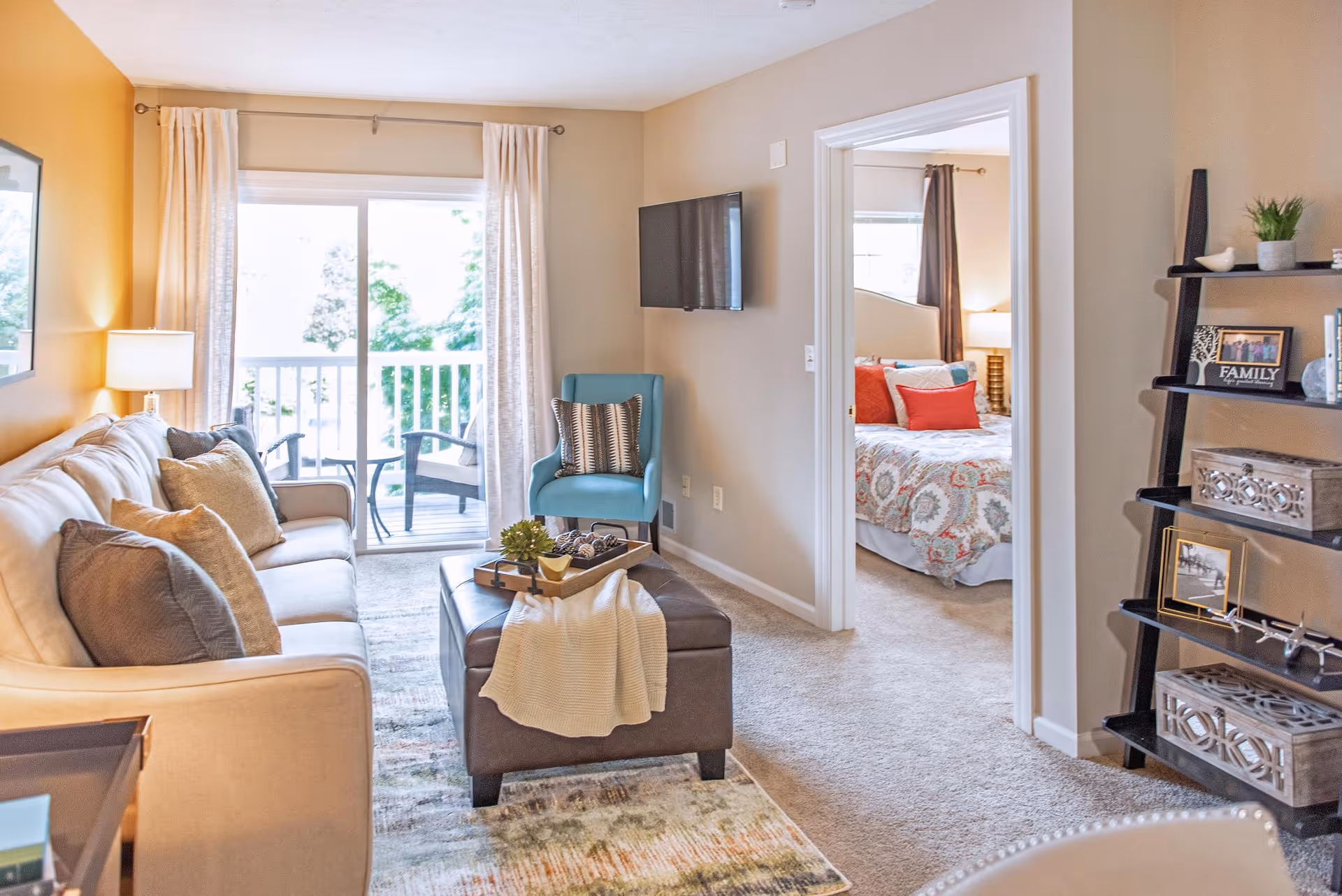 A bright and cozy living room with a beige sofa adorned with multiple cushions, a blue armchair, and a dark ottoman with a tray and a light-colored throw blanket. The room has a sliding glass door leading to a balcony with outdoor chairs. To the right, an open doorway reveals a bedroom with a bed decorated with patterned bedding and red accent pillows. A black ladder-style shelf with decorative items and framed photos is against the wall.