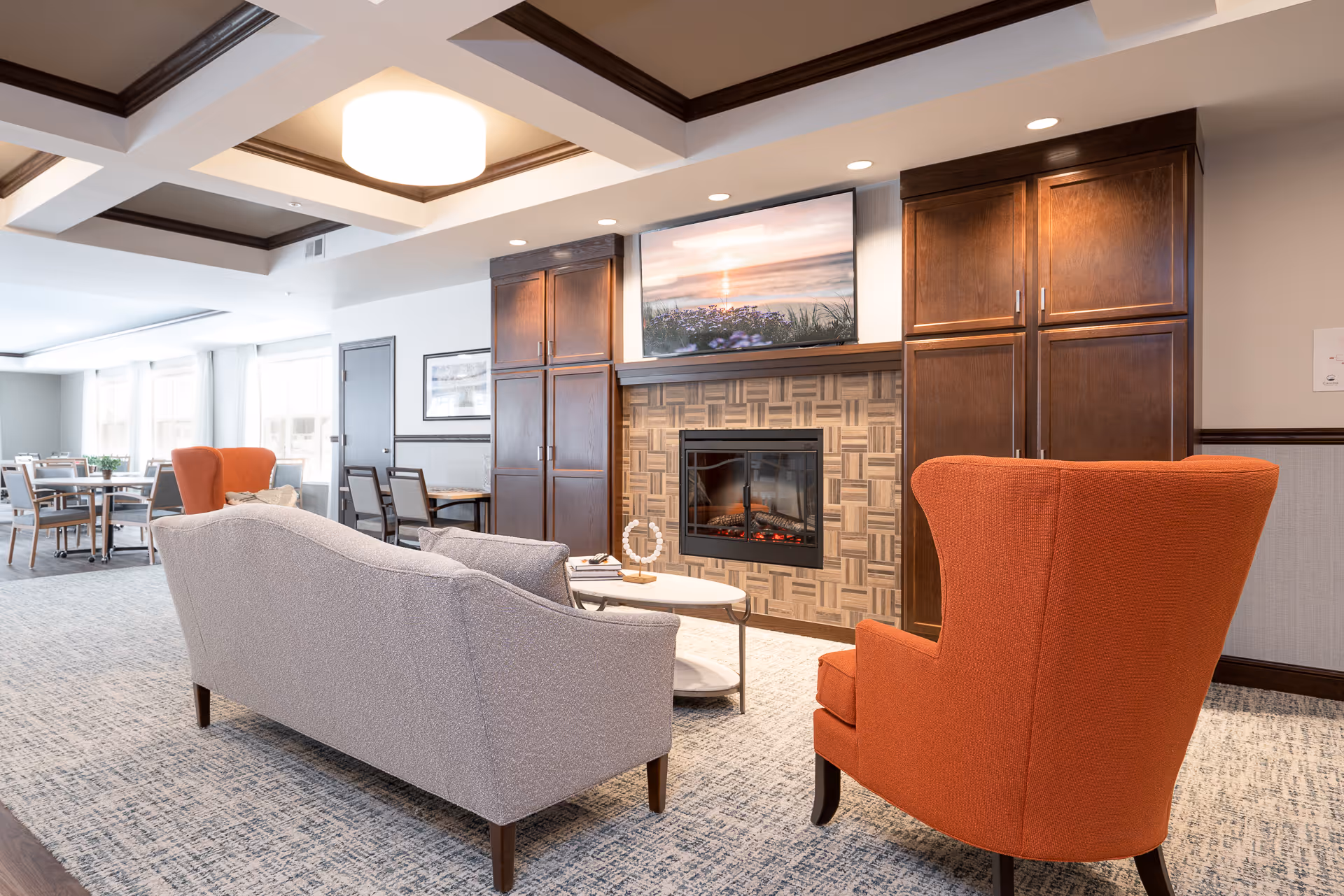 A cozy senior living common area featuring a gray sofa and an orange armchair facing a modern electric fireplace with a TV mounted above it. The room has wooden cabinetry on either side of the fireplace, a patterned carpet, and a ceiling with recessed lighting and a central light fixture. In the background, there are dining tables and chairs near large windows letting in natural light.