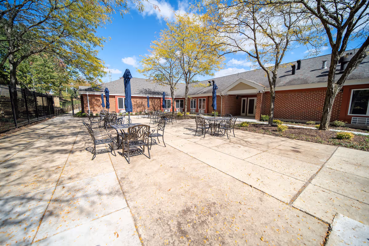 Outdoor patio area at Independence Court of Hyattsville with metal tables and chairs, some tables have closed blue umbrellas. The patio is surrounded by trees with yellowing leaves and a brick building with multiple windows and a covered entrance. The sky is blue with scattered clouds.