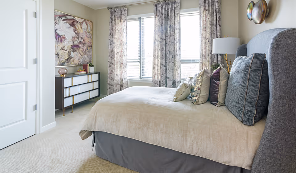 A bedroom with a neatly made bed featuring multiple decorative pillows. The room has light-colored walls, a window with floral curtains, a modern dresser with decorative items on top, and a large abstract painting on the wall.