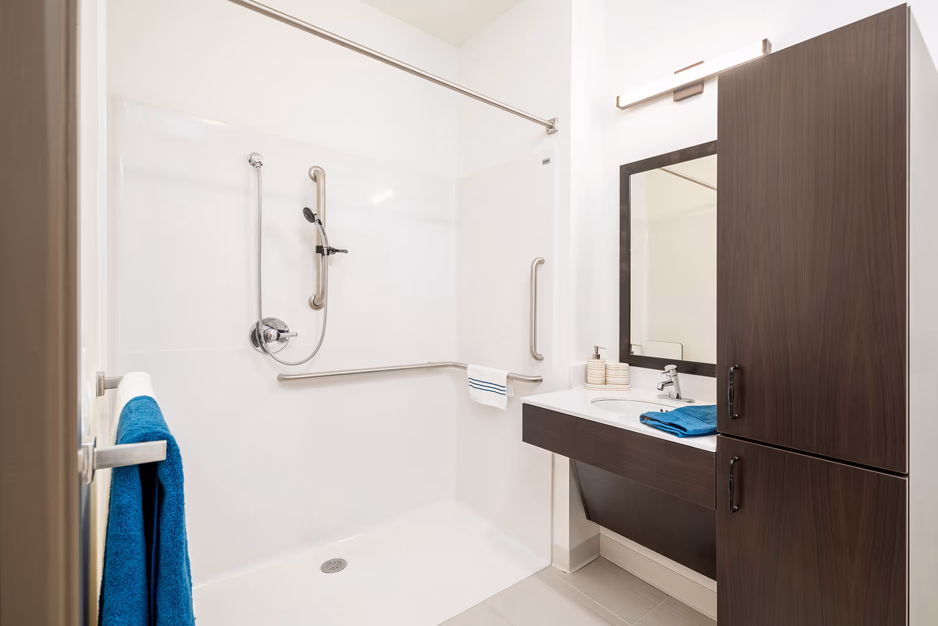 A modern senior living bathroom featuring a walk-in shower with grab bars and a handheld showerhead. The bathroom has a white color scheme with a dark wood vanity cabinet, a mirror, and a sink with a blue towel and soap dispenser on the counter. A blue towel hangs on a towel bar near the shower.