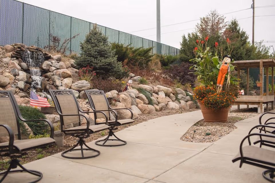 Outdoor seating area with several metal and mesh chairs arranged along a curved concrete pathway. There is a small waterfall feature built with rocks on the left side, surrounded by plants and an American flag. On the right side, there is a large flower pot with orange flowers and a scarecrow decoration. A wooden pergola structure is visible in the background with trees and a green fence behind.