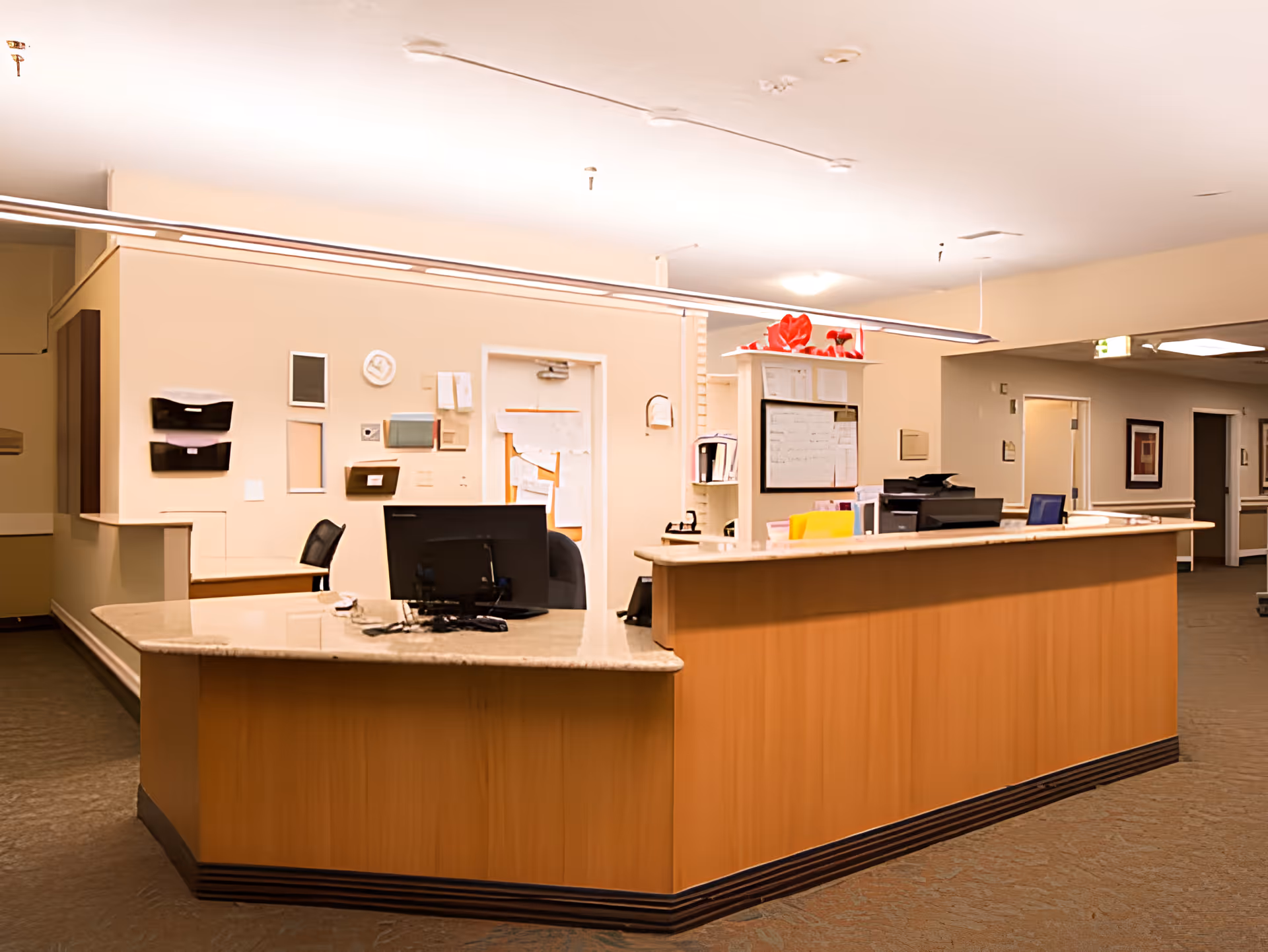 Reception desk area inside a senior living facility with a computer monitor, office chair, and various papers and folders on the counter. The walls are beige with some mounted organizers and a whiteboard. The hallway extends to the right with doors and framed pictures on the walls.