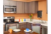 A kitchen area with wooden cabinets, a stainless steel microwave and stove, a sink, and a countertop with a vase of flowers and colorful cone-shaped objects. In the foreground, a dining table is set with plates, bowls, and mugs.