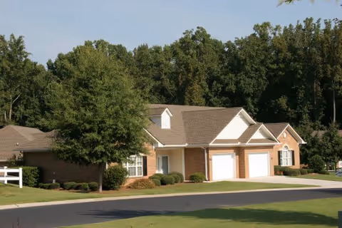 A single-story residential building with brick and white siding exterior, featuring two garage doors, surrounded by green lawns and trees under a clear sky.