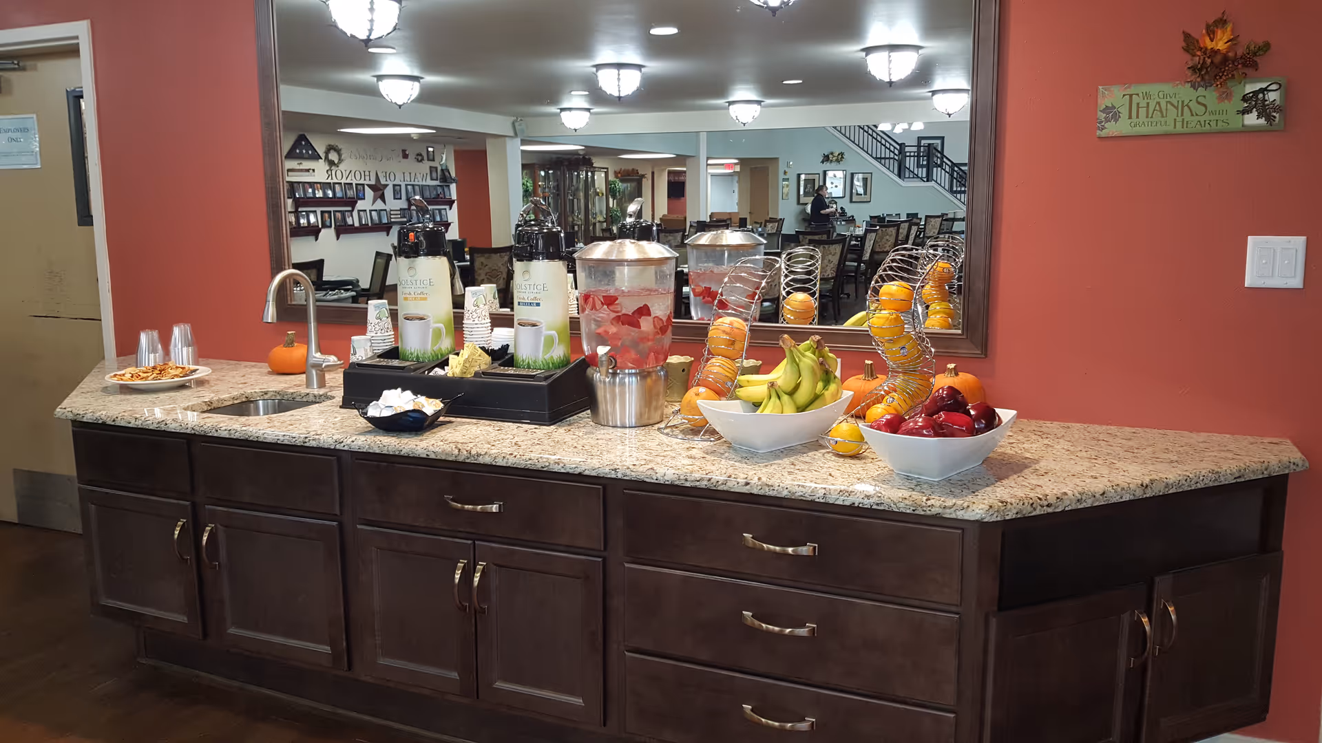 Granite buffet counter in a communal dining area with bowls of fruit, beverage dispensers, a sink, and a large mirror reflecting dining tables.