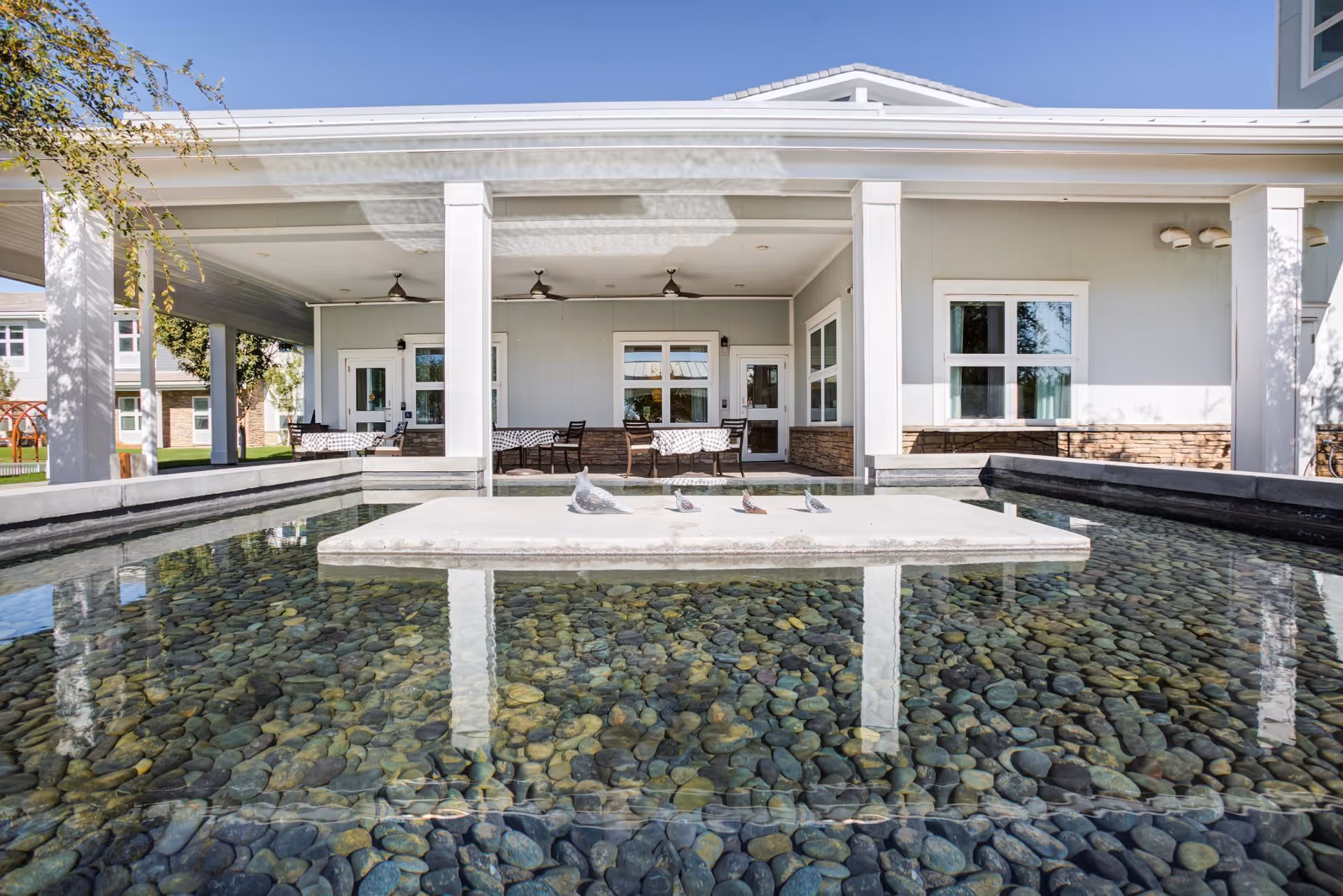 Decorative reflecting pool filled with river stones and a central platform with bird sculptures in front of a covered patio and building façade.