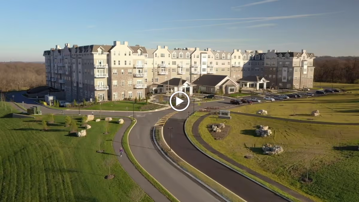 A large multi-story retirement living facility building with beige and light brown exterior walls, surrounded by well-maintained green lawns and a curved driveway with parked cars. The sky is clear and blue.