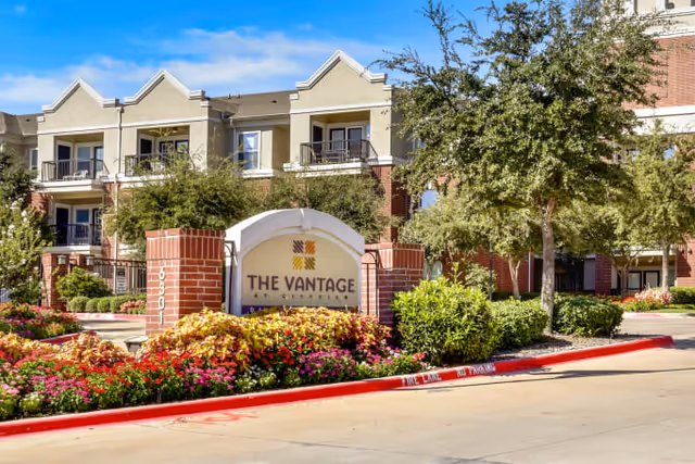 Exterior view of The Vantage at Cityview senior living facility with a prominent sign in front surrounded by colorful flowers and greenery. The building behind has multiple balconies and a mix of brick and beige siding under a clear blue sky.