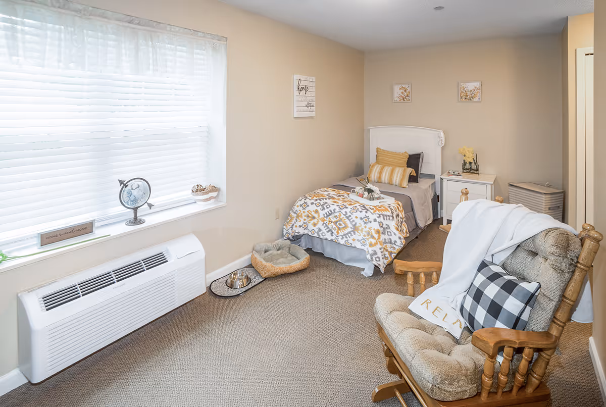 A cozy bedroom with a single bed covered in a patterned blanket and pillows. Next to the bed is a white nightstand with a small vase of flowers and a pair of glasses. A cushioned rocking chair with a black and white checkered pillow and a white blanket is in the foreground. A window with white blinds lets in natural light, and a small globe and decorative items sit on the windowsill. A pet bed and food bowl are on the floor near the bed. The walls are beige with framed artwork and a decorative sign.