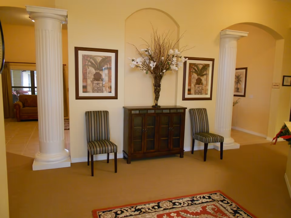 Entryway sitting area with two striped chairs flanking a wooden cabinet topped by a tall floral arrangement, framed artwork, and two decorative columns.