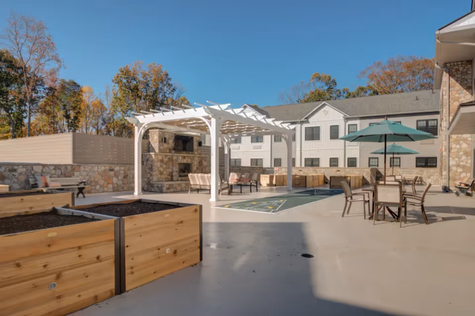 Outdoor patio area at Mercer Hill at Doylestown featuring raised garden beds, a white pergola with seating underneath, a stone fireplace, shuffleboard court, and tables with umbrellas. The building with multiple windows is visible in the background along with trees under a clear blue sky.