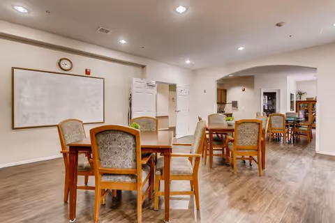 Communal dining/activity room with wooden tables and upholstered chairs on wood flooring, a whiteboard and clock on the wall, and an arched doorway leading to more seating.