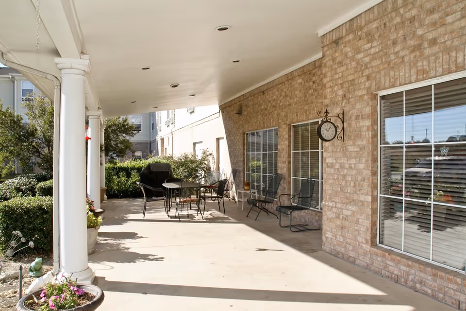 Covered outdoor patio with white columns, brick exterior wall, windows, a decorative clock, and patio tables and chairs.