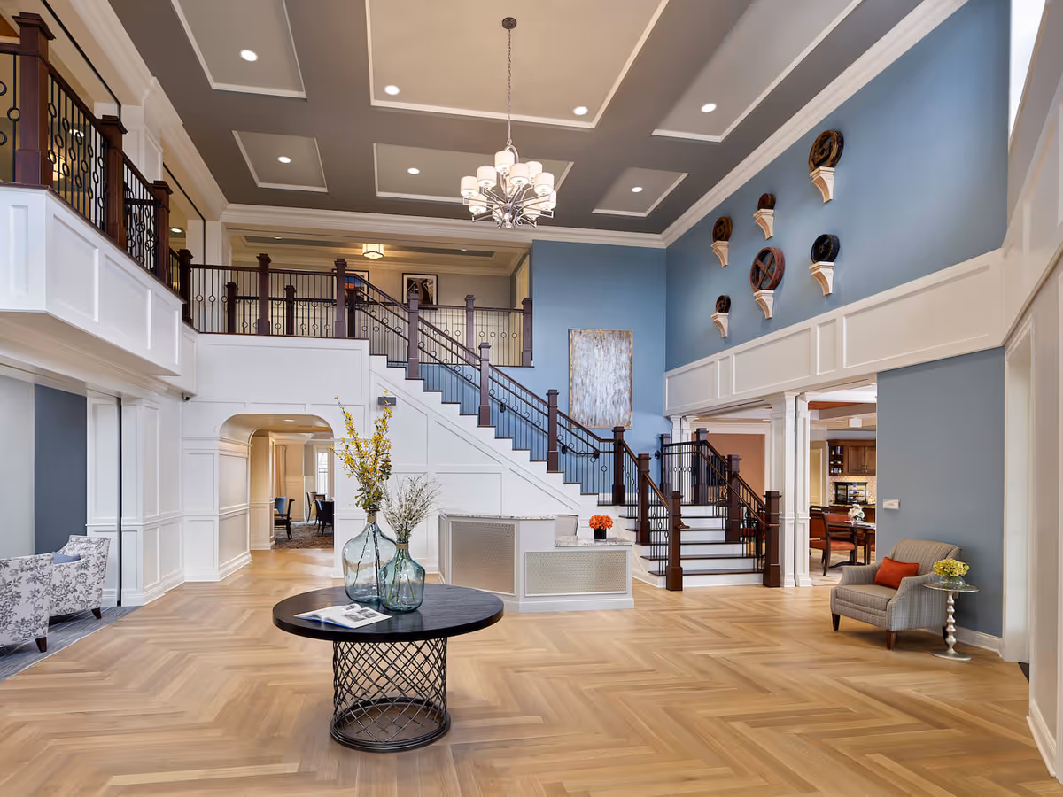 Spacious and elegant interior of a senior living facility with a high ceiling, a chandelier, a staircase with dark wood railings, blue and white walls, a round table with decorative vases and flowers, and comfortable seating areas.