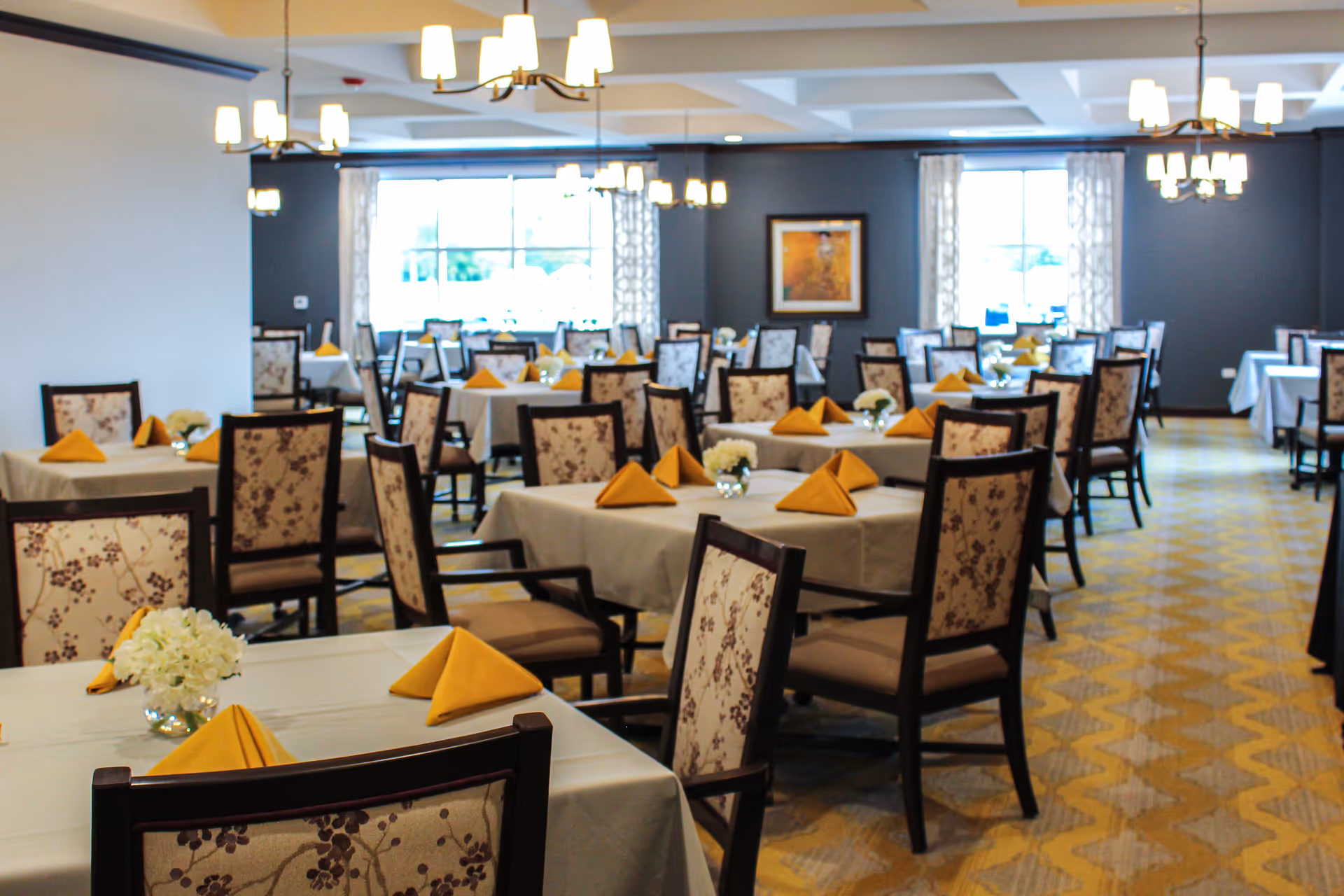 Spacious dining room with tables set with folded yellow napkins, floral centerpieces, and patterned chairs.