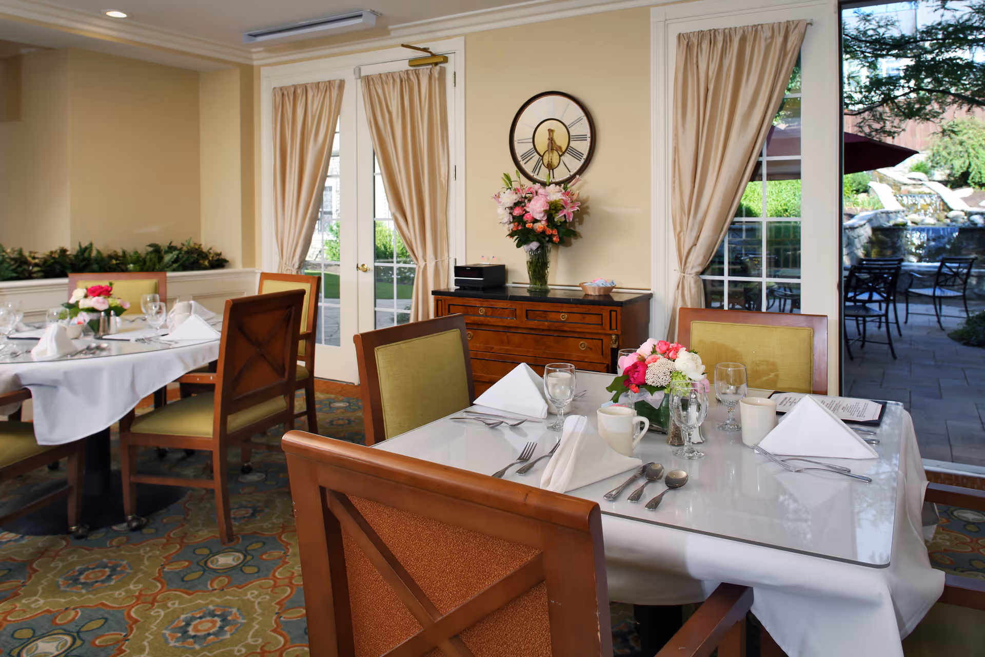 A dining room with tables set for a meal, featuring white tablecloths, folded napkins, glassware, and floral centerpieces. The room has wooden chairs with green upholstery, beige walls, and large windows with beige curtains that look out onto a patio with outdoor seating and a water feature.