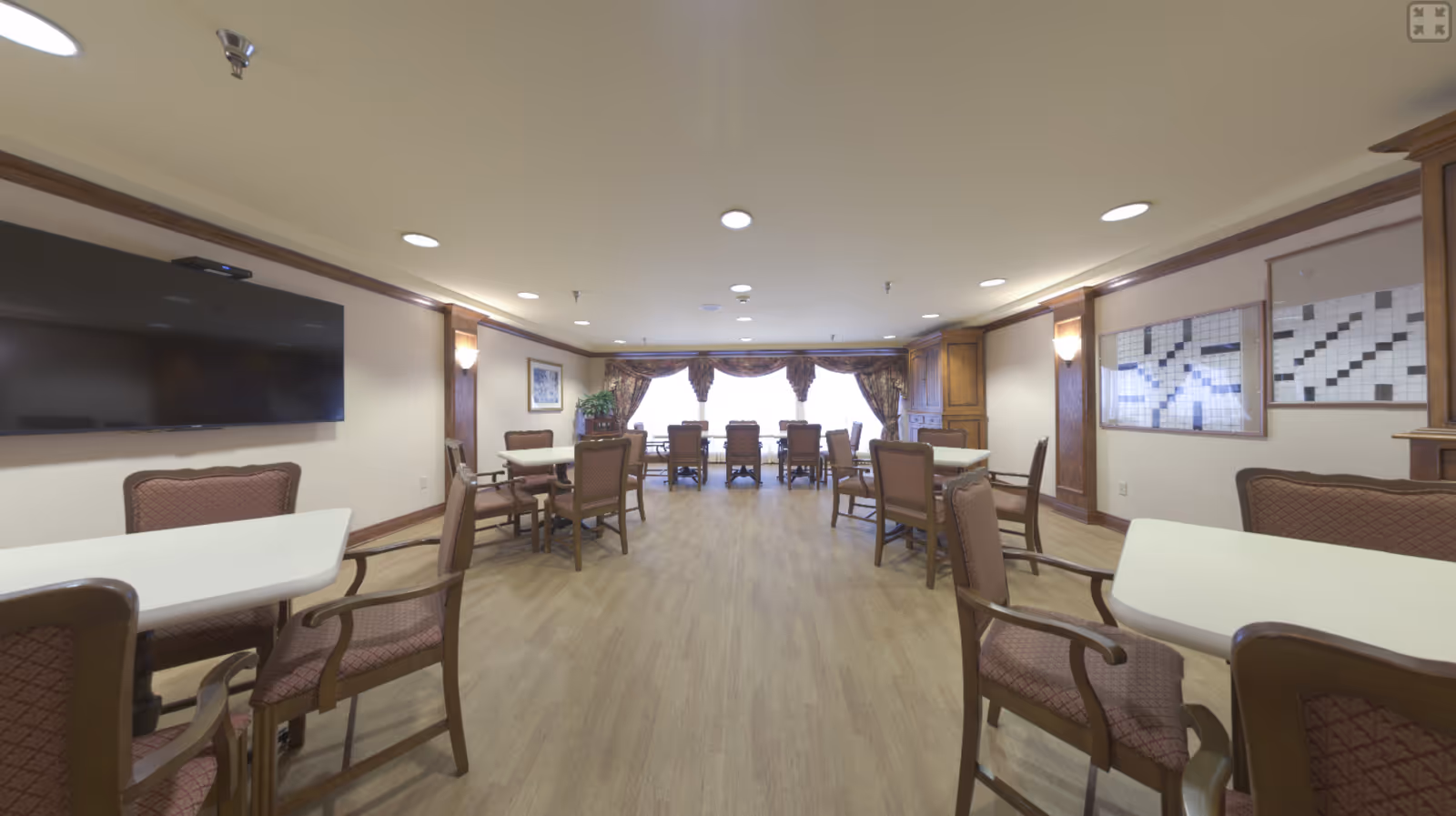 A spacious dining/activity room with multiple tables and upholstered chairs, a wall-mounted TV, and windows with drapes at the far end.