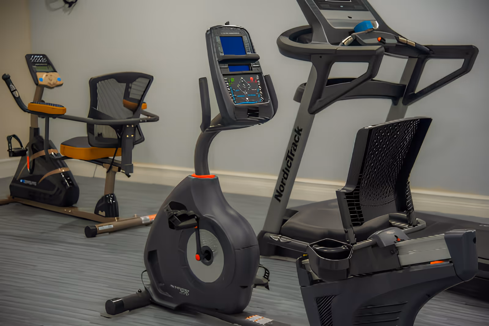 Indoor fitness room with exercise equipment including a recumbent bike, an upright stationary bike, and a treadmill against a plain wall with gray flooring.