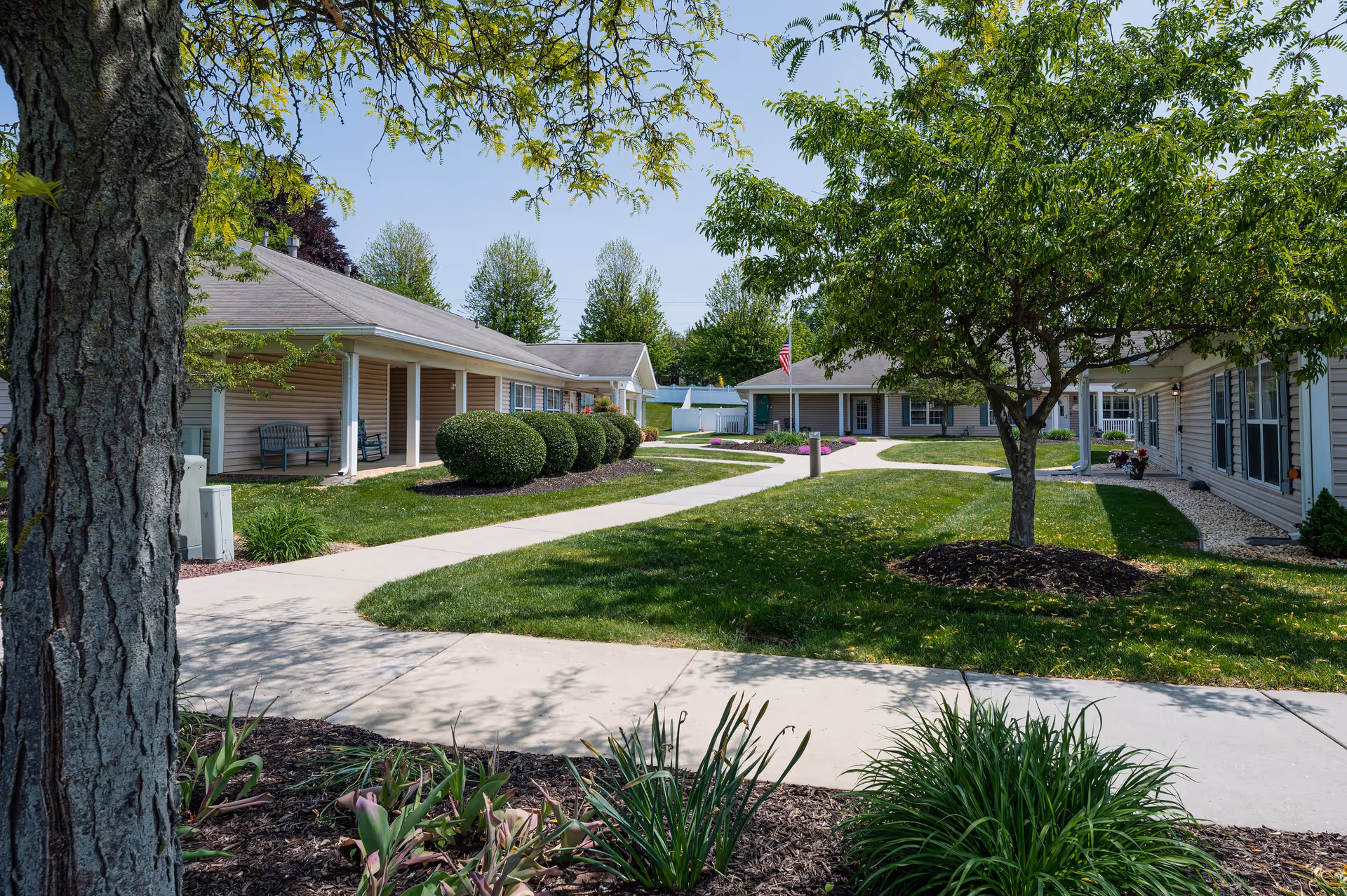 A landscaped outdoor courtyard area at Linden Village A ProMedica Senior Living Community featuring a paved walkway, green grass, trimmed bushes, trees, and single-story buildings with covered porches and benches.