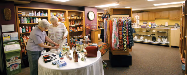 Two elderly women browsing items on a round table in a small retail shop area within a retirement community. The shop has shelves stocked with various products including snacks, toiletries, and jewelry, as well as racks with colorful scarves and accessories. The setting is warm and inviting with carpeted floors and wooden shelving.