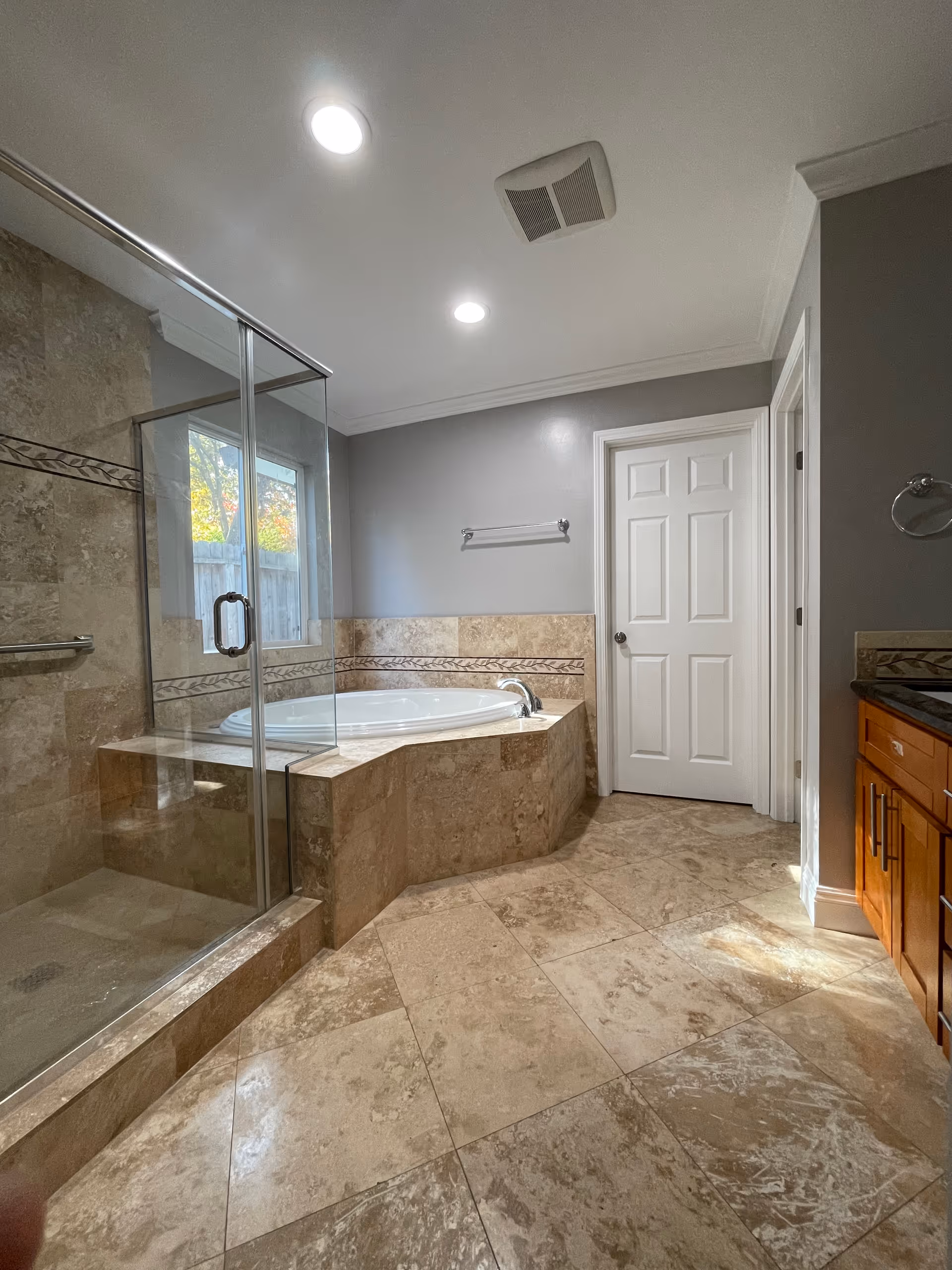 A spacious bathroom featuring a large corner bathtub with beige tile surround and decorative border, a glass-enclosed shower with matching tile, a white door, and wooden cabinetry with a granite countertop. The walls are painted gray and the floor is covered with beige tiles.