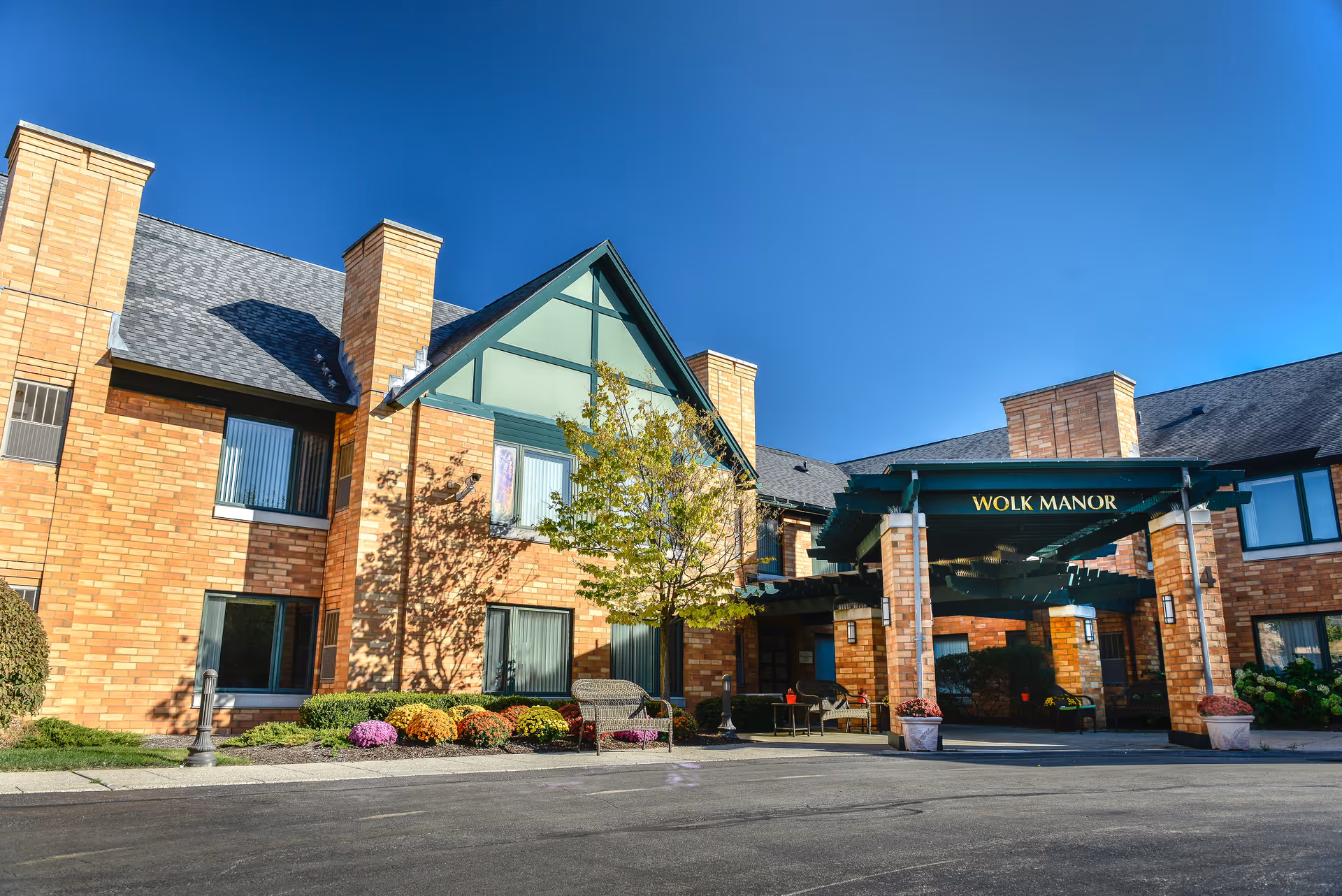 Exterior view of Wolk Manor Assisted Living facility showing a brick building with multiple windows, a covered entrance with green beams, a tree, benches, and colorful flower bushes under a clear blue sky.