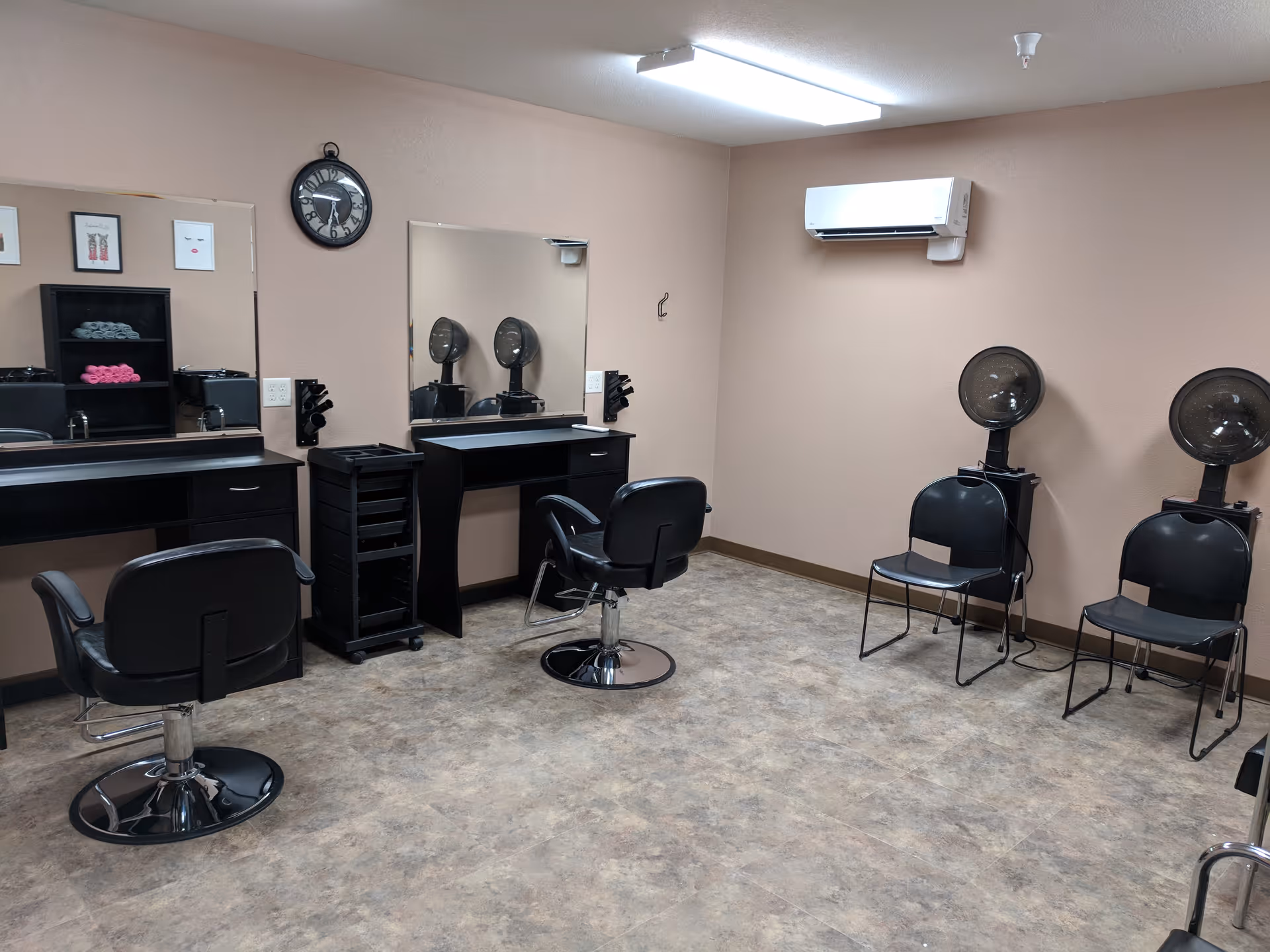 Interior view of a hair salon area in an assisted living facility featuring two black styling chairs in front of large mirrors, a black rolling cart, shelves with towels, two black chairs with hair dryers attached, beige walls, and a wall-mounted air conditioning unit.