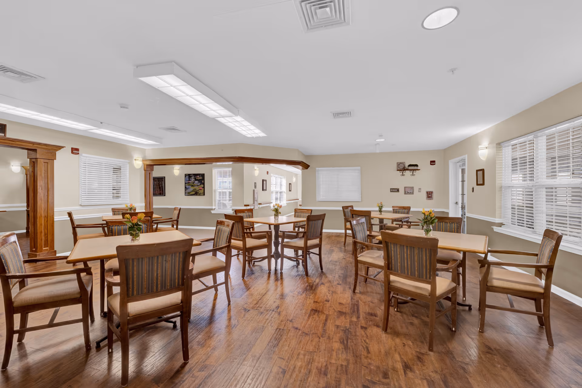 A bright and spacious dining room with several wooden tables and chairs arranged neatly. Each table has a small vase with flowers. The room has wooden flooring, beige walls with white trim, and multiple windows with white blinds allowing natural light to enter. Ceiling lights and wall sconces provide additional lighting.