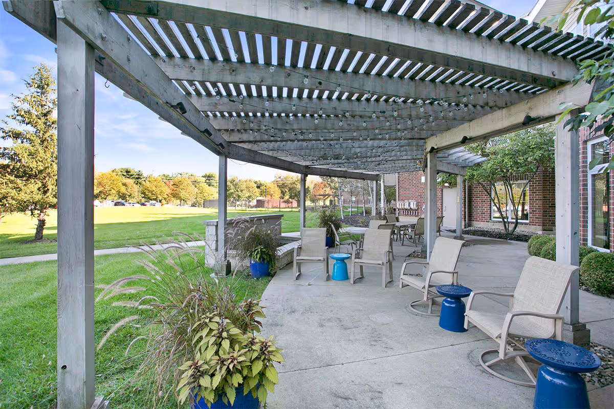 Outdoor patio area at Walnut Ridge with a wooden pergola overhead, several beige chairs with blue side tables, potted plants, and a view of a grassy lawn and trees in the background.