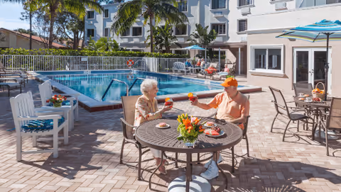 An elderly couple sitting at a round outdoor table by a swimming pool, toasting with drinks. The pool area is surrounded by lounge chairs, umbrellas, and palm trees, with a multi-story building in the background.