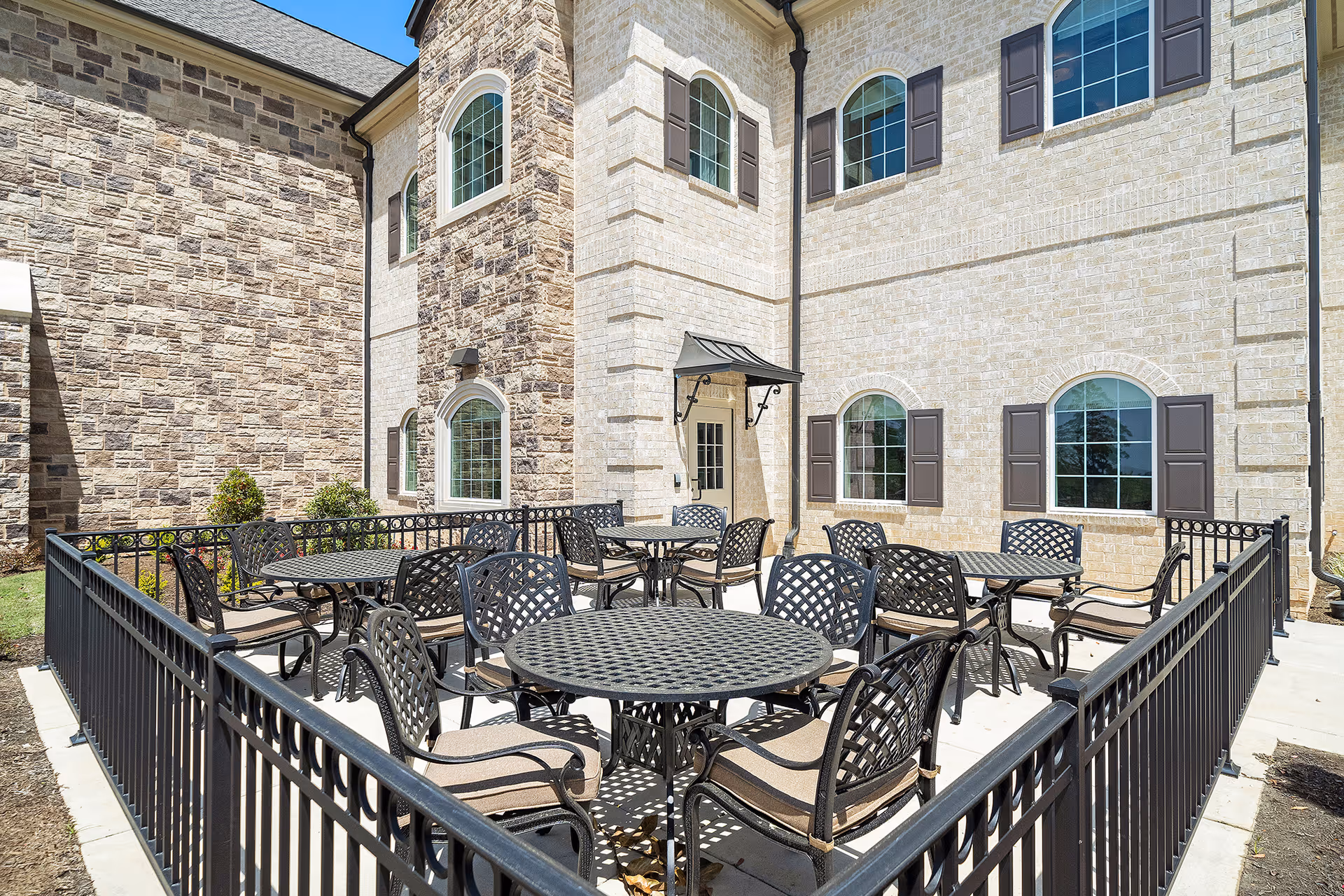 Outdoor patio area at Worthington Manor with multiple round metal tables and cushioned chairs surrounded by a black metal fence. The patio is adjacent to a beige brick building with arched windows and brown shutters under a clear blue sky.
