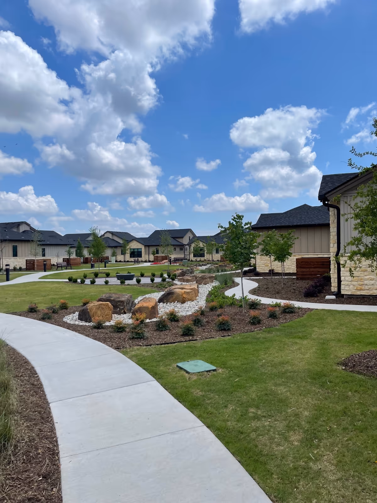 A landscaped outdoor area with a curved concrete walkway, green grass, small trees, shrubs, and large decorative rocks. Several single-story buildings with stone and beige siding are visible under a partly cloudy blue sky.
