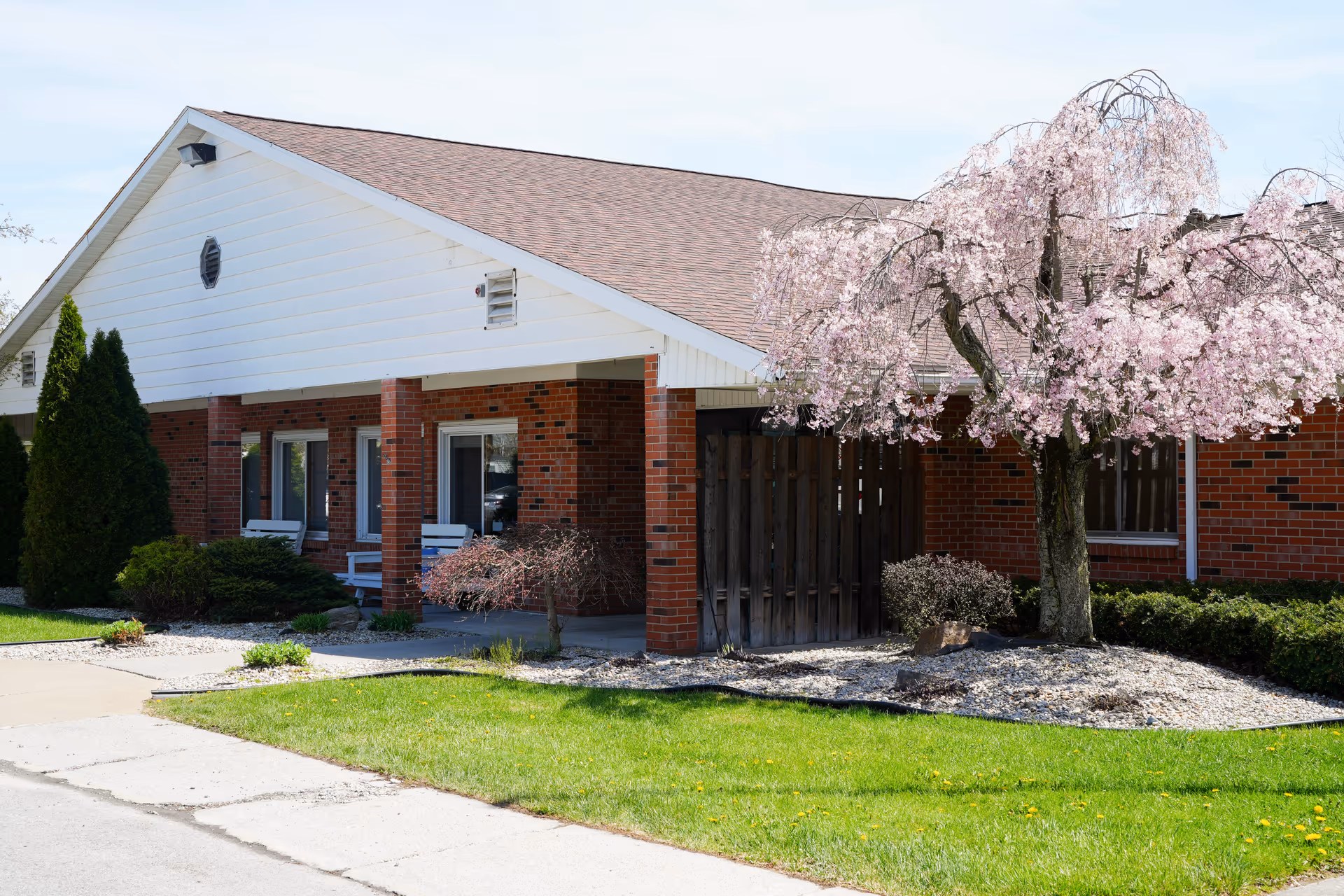Front exterior of a single-story brick senior living building with a covered porch, white rocking chairs, and a blooming pink tree.