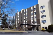 Multi-story beige and brown senior living facility with a covered entrance, driveway, and lawn under a clear blue sky.
