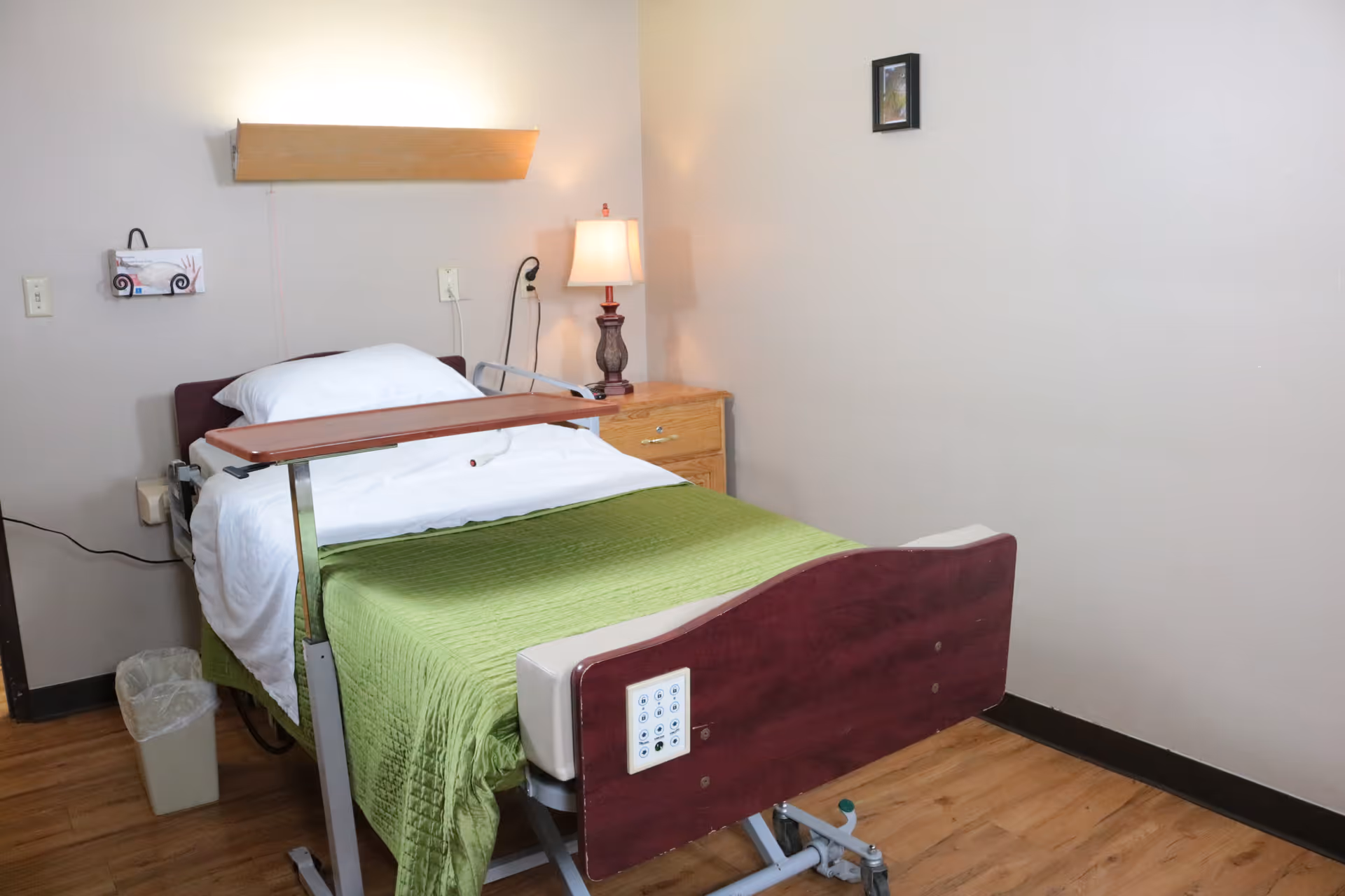 A simple room with a hospital-style bed covered with a green blanket and white sheets. There is an adjustable overbed table positioned over the bed. Next to the bed is a wooden nightstand with a lamp on it. The walls are plain with a small framed picture hanging above the nightstand. The floor is wooden, and there is a small trash can beside the bed.