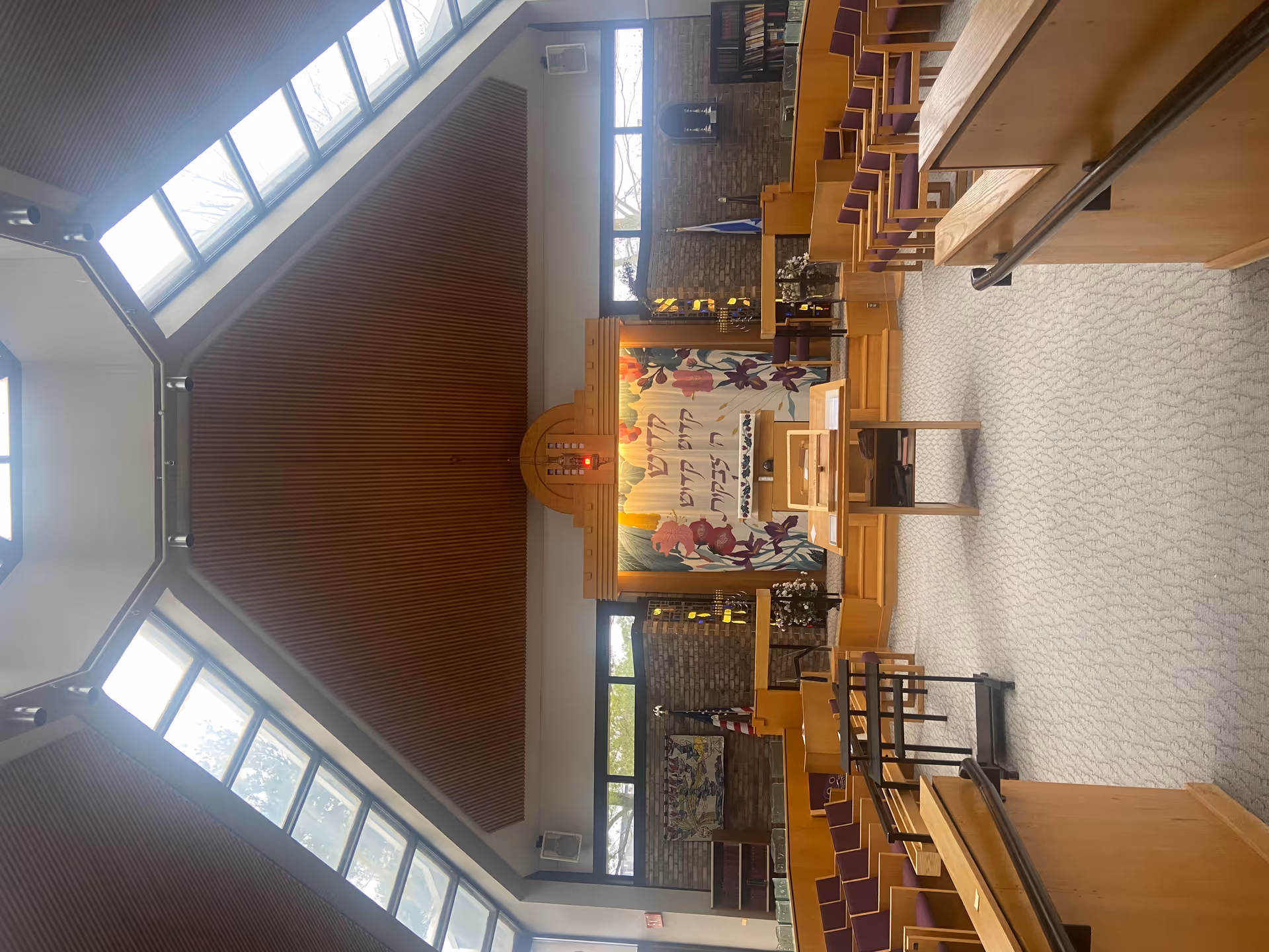 Interior view of a synagogue or chapel with wooden pews and chairs arranged facing a raised platform with a lectern and an ark covered by a decorative curtain with Hebrew text and floral designs. The ceiling is high with large skylights allowing natural light to enter.