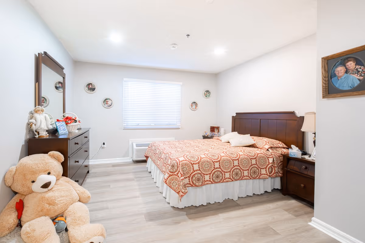 A cozy bedroom with a large bed covered in a red and beige patterned bedspread, two pillows, and a wooden headboard. To the right of the bed is a wooden nightstand with a lamp and tissues. On the left side of the room is a dark wooden dresser with a mirror above it, decorated with a doll, a basket of flowers, and a framed photo. A large teddy bear sits on the floor near the dresser. The walls are light-colored with small decorative plates and a framed photo of an elderly couple. The floor is light wood, and a window with blinds is centered on the back wall.