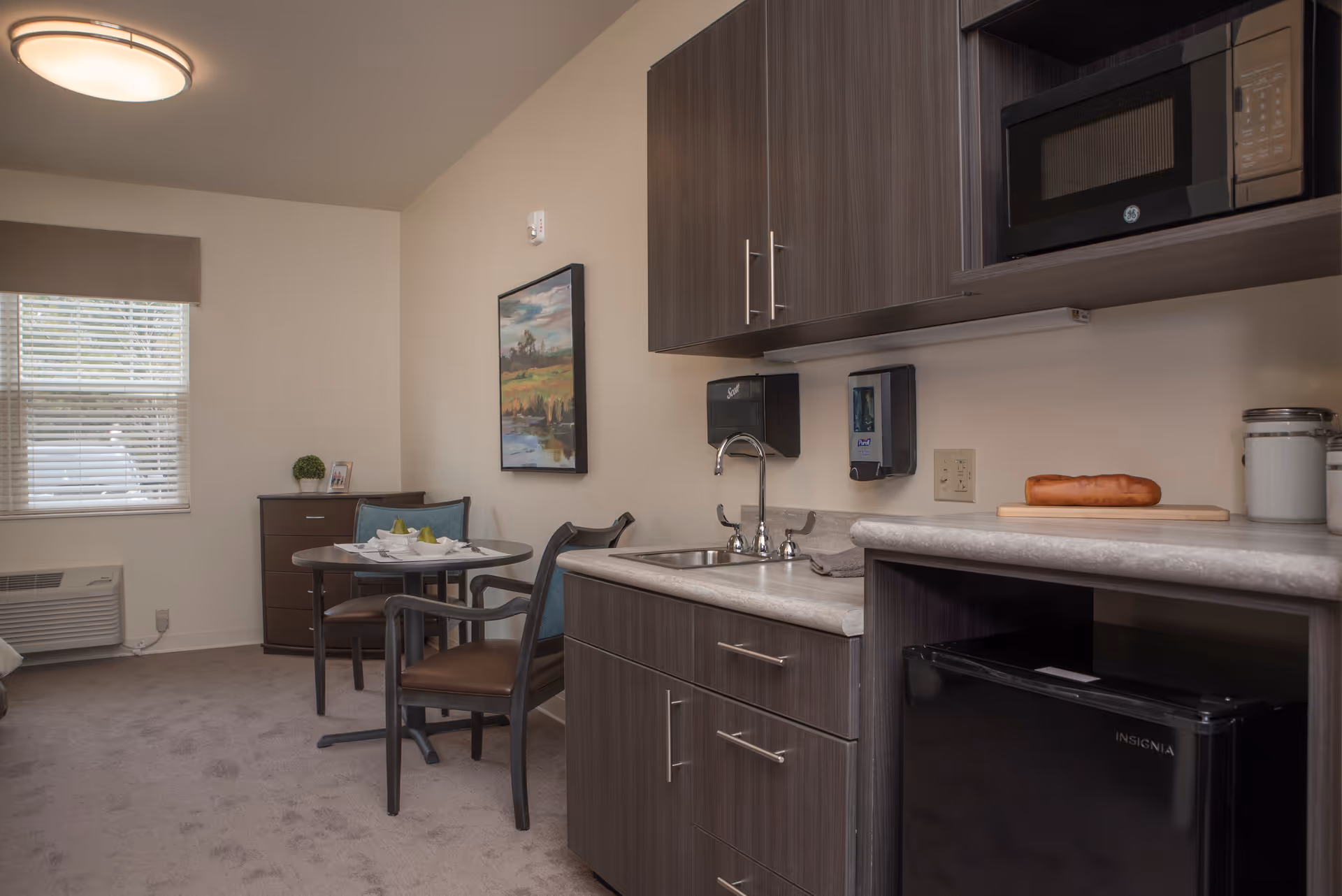 Small studio kitchenette and dining area with a sink, microwave, mini-fridge, and a round table by a window.