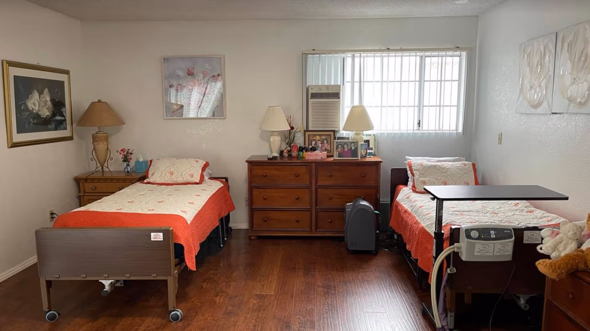 A bedroom with two single beds, each covered with white and red bedding. Between the beds is a wooden dresser with two lamps and framed photos on top. The room has wooden flooring, a window with vertical blinds, and wall art above each bed. One bed has a medical device attached to it.