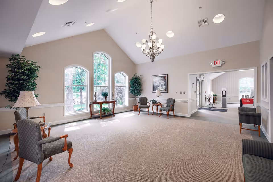 A spacious, well-lit common area with beige walls and carpeted floor. The room features three large arched windows letting in natural light, two potted plants, a chandelier hanging from the vaulted ceiling, and several upholstered chairs arranged around small wooden tables. There is a framed picture on the wall and an exit sign above a doorway leading to another room.