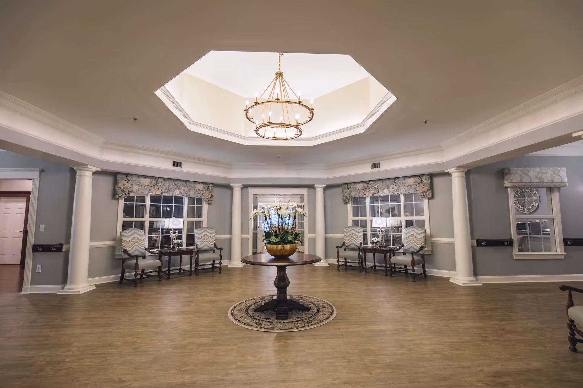 A spacious, elegant common area with a round wooden table in the center holding a large potted plant with white flowers. The room features a high octagonal ceiling with a large chandelier, light gray walls with white trim, and several windows with floral valances. There are pairs of cushioned chairs and small tables arranged symmetrically along the walls, and white columns supporting the ceiling structure.