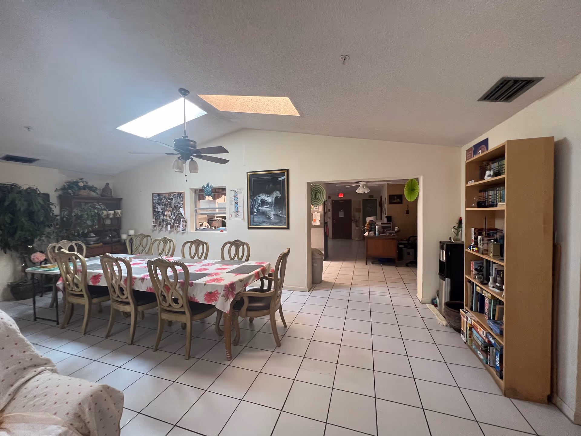 A spacious dining area with a long table covered by a floral tablecloth and surrounded by eight chairs. The room has white tiled floors, a ceiling fan with lights, and two skylights. There are plants and a wooden cabinet with decorative items on the left side, a bookshelf filled with books on the right, and a doorway leading to another room with office furniture and a red exit sign in the background.