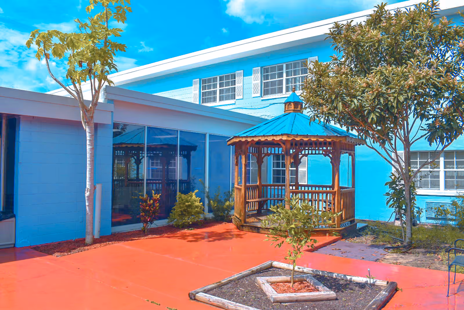 Outdoor courtyard area at Haines Manor Assisted Living featuring a wooden gazebo with a blue roof, surrounded by small trees and plants, with a bright red paved ground and a light blue building in the background.