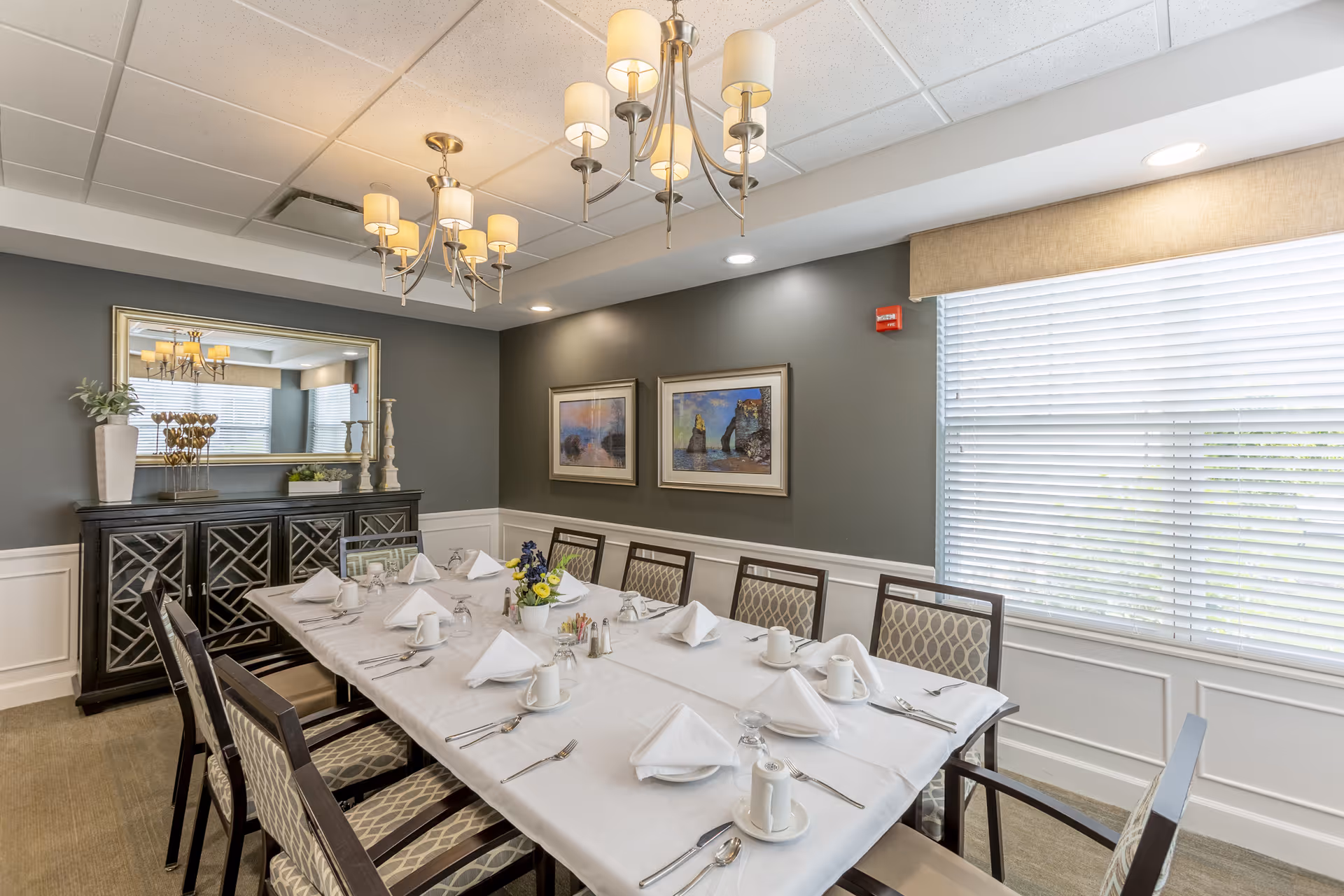 A formal dining room with a long rectangular table set for a meal. The table is covered with a white tablecloth and has white napkins, cups, and silverware neatly arranged. There are ten upholstered chairs around the table. The room features gray walls with white wainscoting, two framed landscape paintings, a large mirror above a dark wooden sideboard, and two modern chandeliers hanging from the ceiling. A large window with white blinds allows natural light into the room.