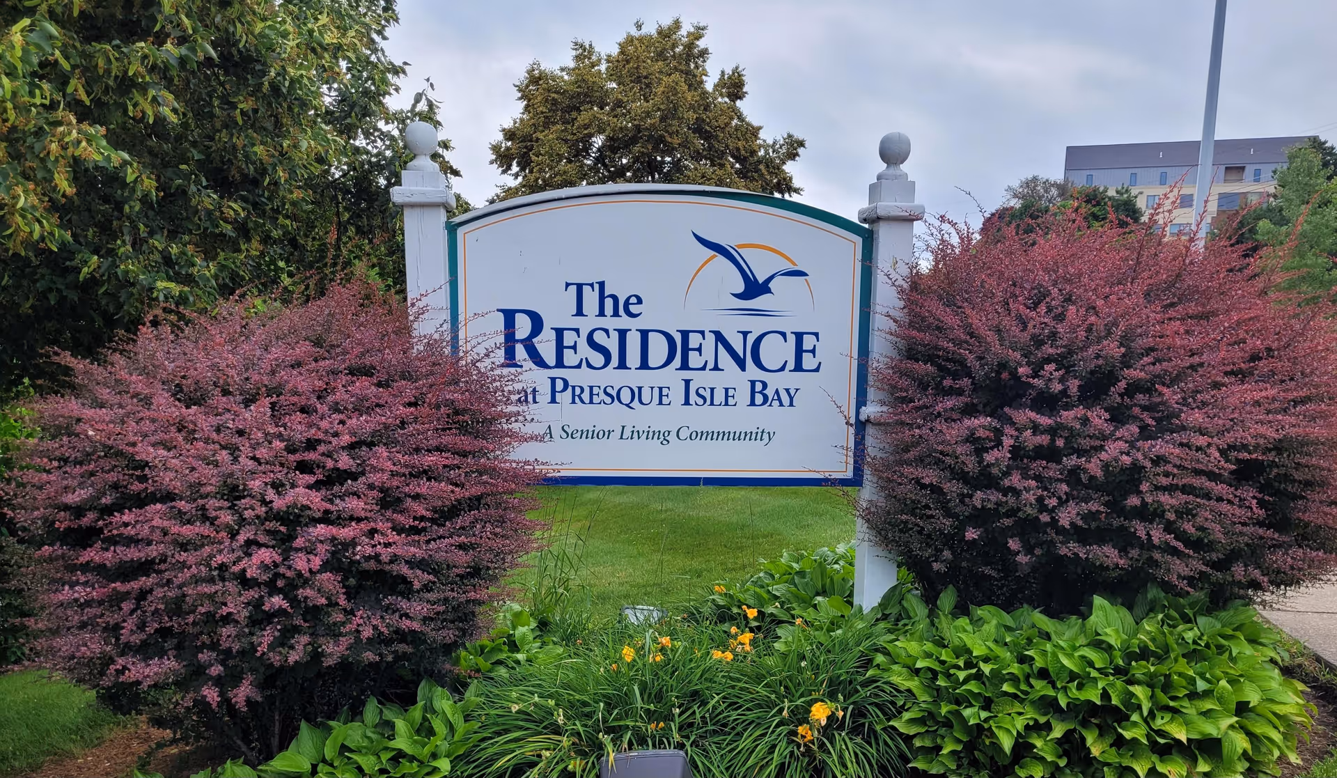 Outdoor sign for The Residence at Presque Isle Bay, a senior living community, surrounded by green grass, purple bushes, and other greenery with a building and cloudy sky in the background.