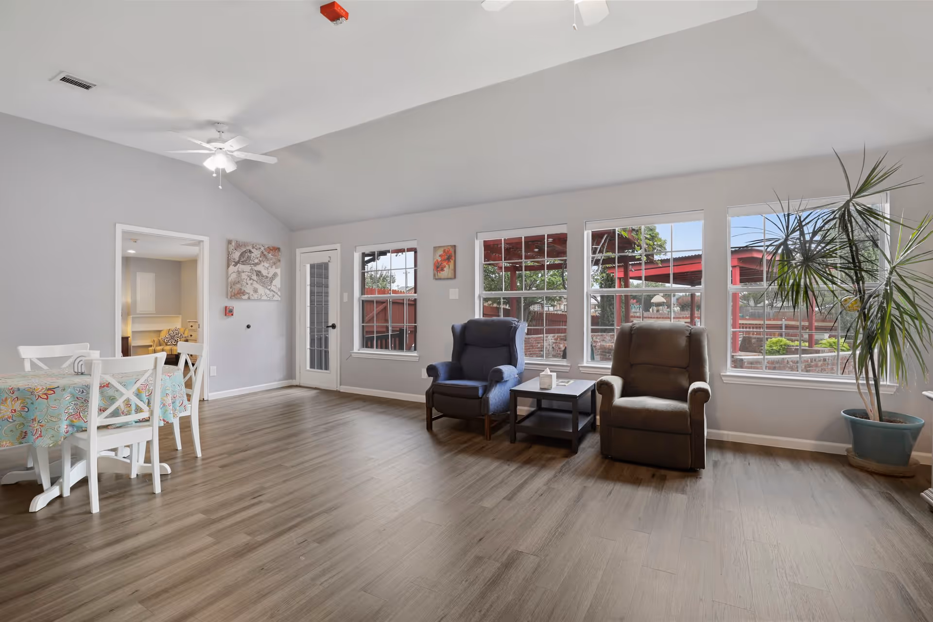 A bright and spacious living area with two armchairs, a small side table, large windows showing an outdoor view, a ceiling fan, a dining table with chairs, and a potted plant in the corner.